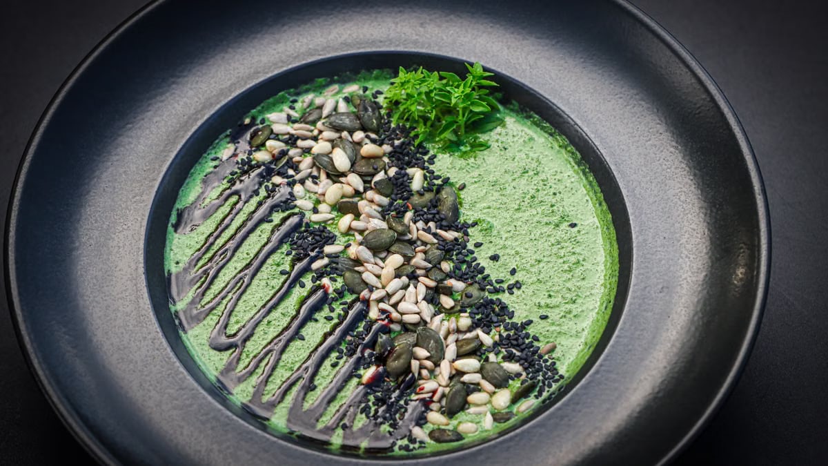 A gourmet green soup served in a black bowl, topped with mixed seeds and drizzled sauce.